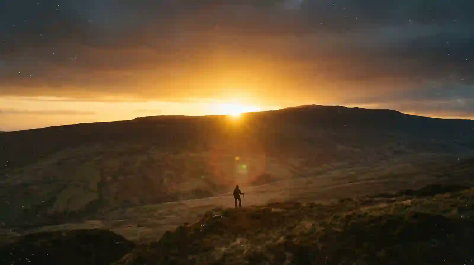 A figure standing on a ridge watching the sun rise over distant mountains